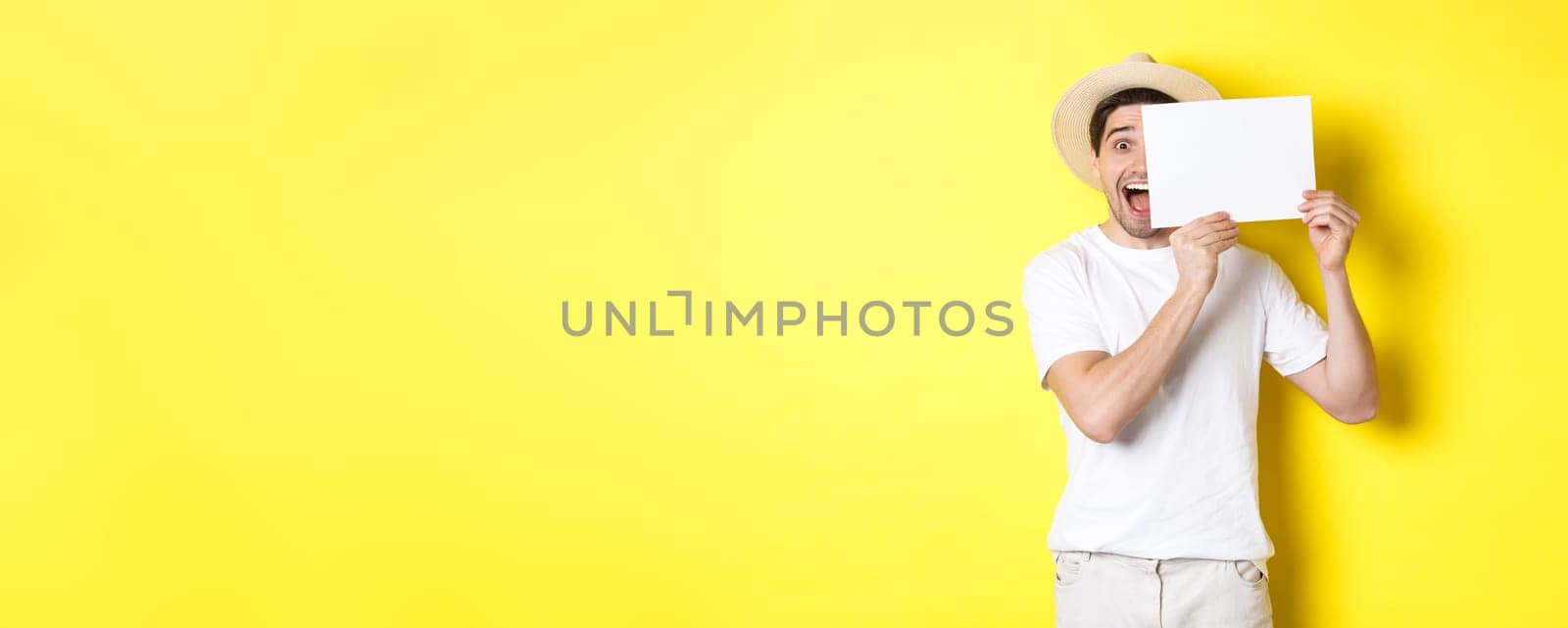 Excited tourist on vacation showing blank piece of paper for your logo, holding sign near face and smiling, standing against yellow background by Benzoix