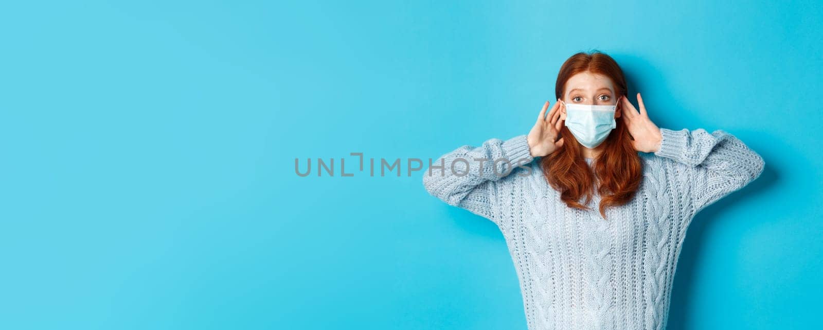 Winter, covid-19 and social distancing concept. Intrigued redhead girl in face mask, eavesdropping, holding hands near ears and listen closer, standing against blue background.
