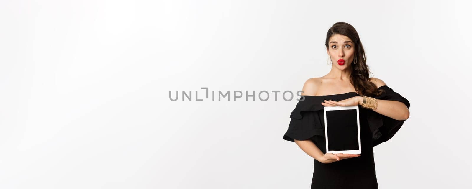Fashion and shopping concept. Beautiful woman with red lipsticks, black dress, showing tablet screen and looking excited, standing over white background by Benzoix