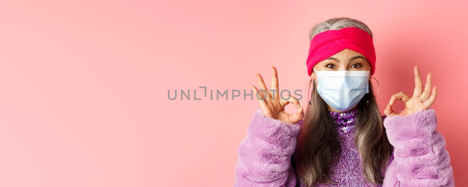 Covid-19, virus and fashion concept. Close-up of fashionable asian senior woman in face mask, showing OK signs and looking happy, pink background.