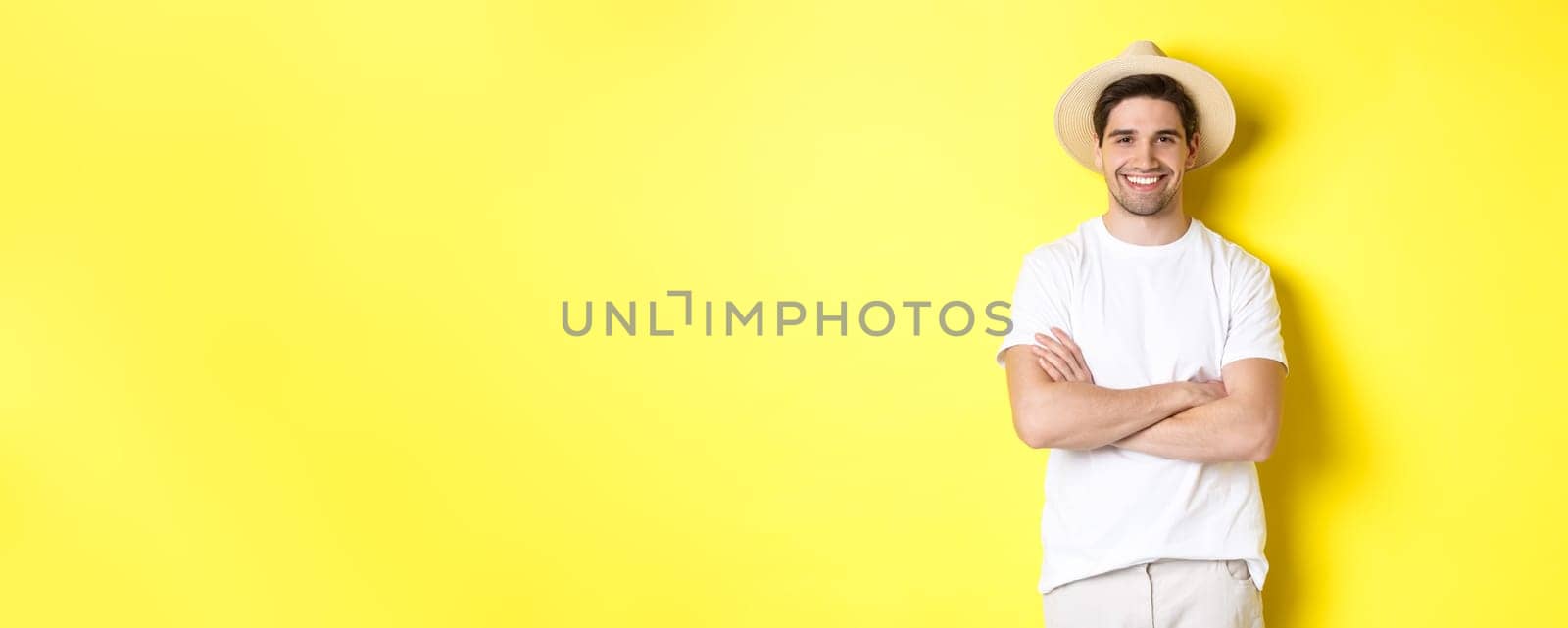 Tourism. Handsome young man looking happy, wearing straw hat for travelling, cross hands on chest and smiling, standing over yellow background by Benzoix