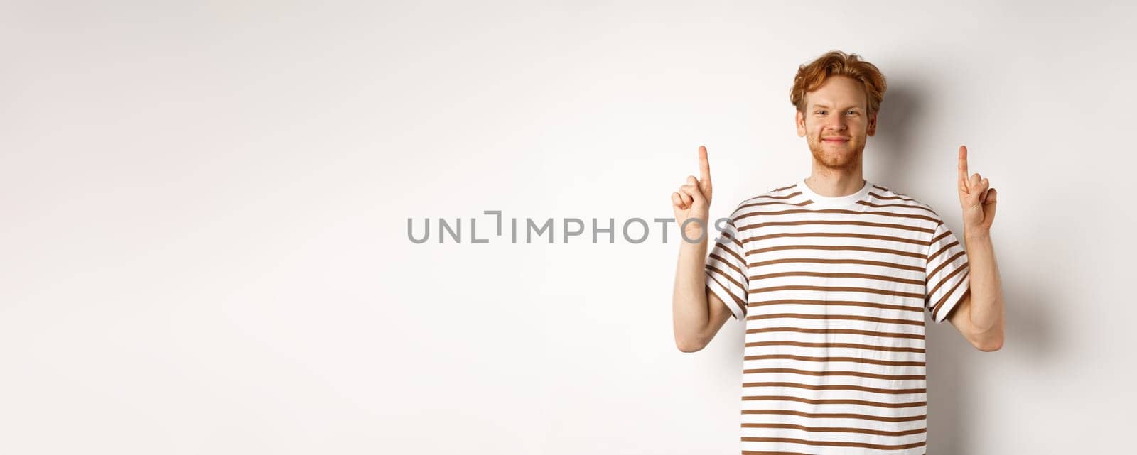Smiling redhead guy with beard, pointing fingers up and showing advertisement, standing over white background.