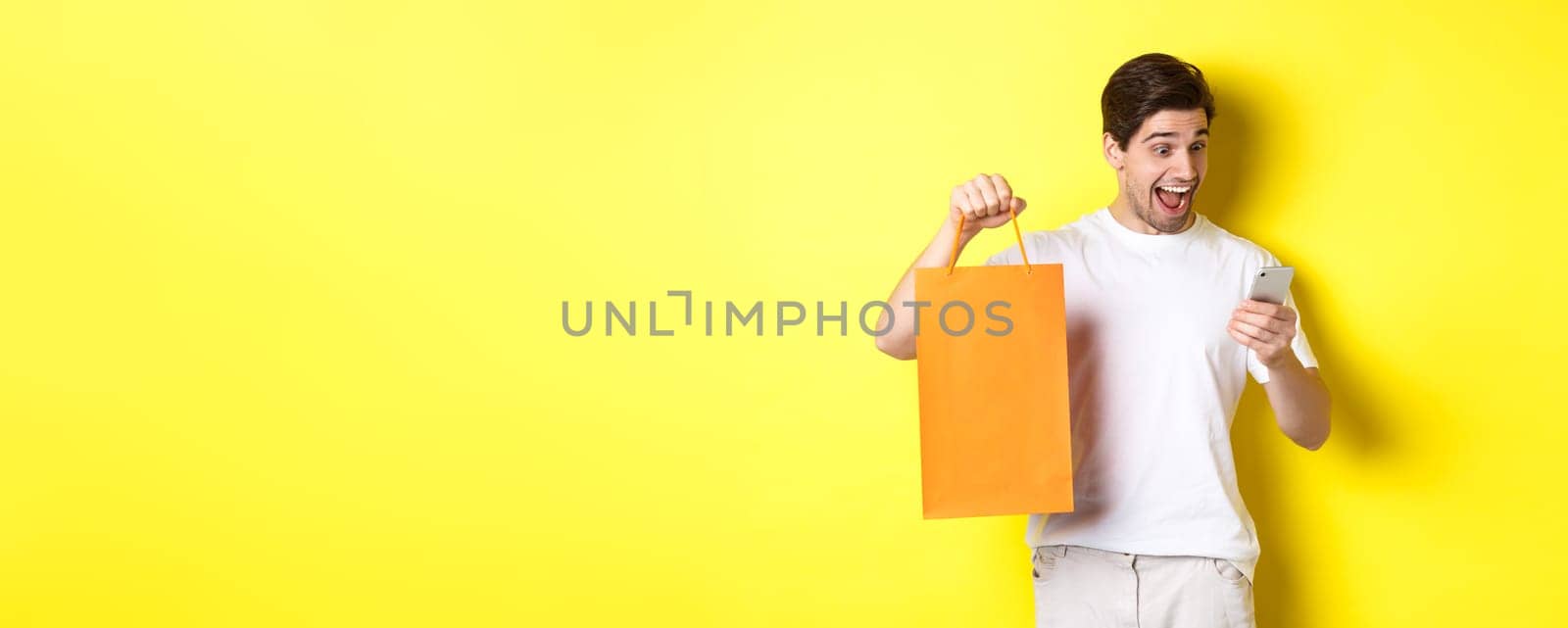 Concept of discounts, online banking and cashback. Surprised man showing shopping bag and looking happy at mobile screen, standing against yellow background.