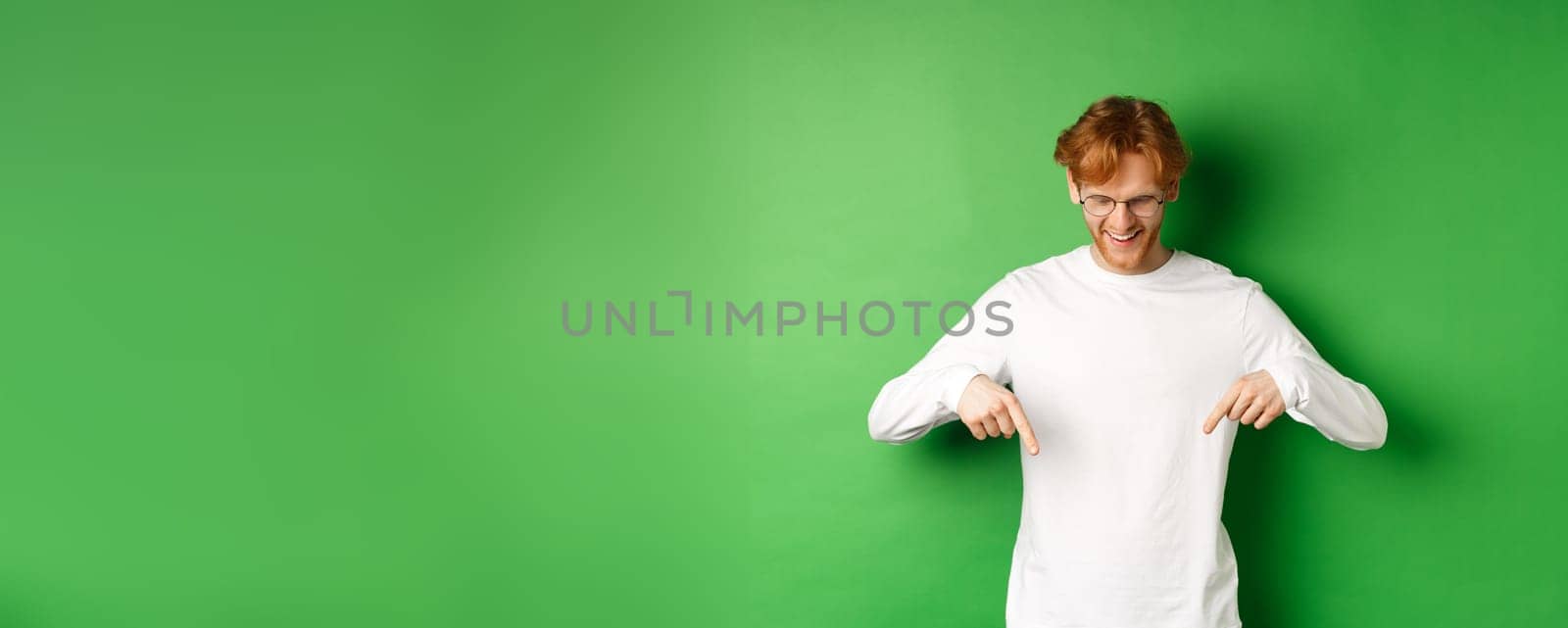Handsome redhead man in glasses and white long-sleeve checking out advertisement, pointing fingers and looking down with pleased smile, standing over green background by Benzoix