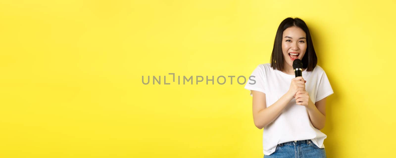 Beautiful asian woman singing karaoke, perform with microphone, smiling happy and looking at camera, standing over yellow background by Benzoix