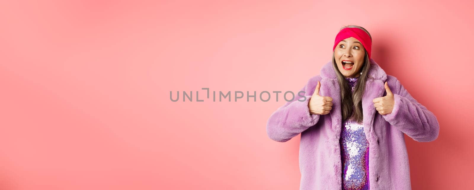 Fashion and shopping concept. Happy asian senior woman in stylish clothes checking out awesome advertisement, looking at upper left corner and showing thumbs-up in approval by Benzoix