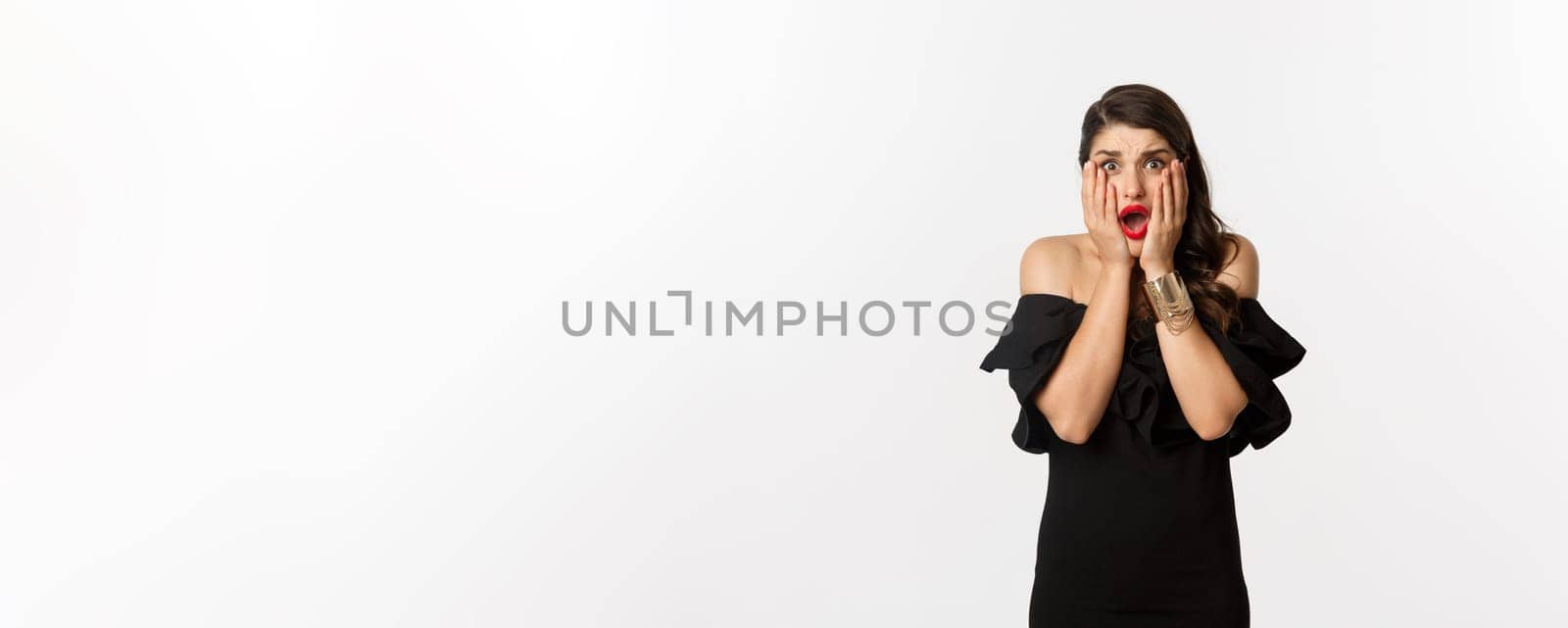 Fashion and beauty. Shocked and worried woman in black dress looking scared, standing anxious over white background.