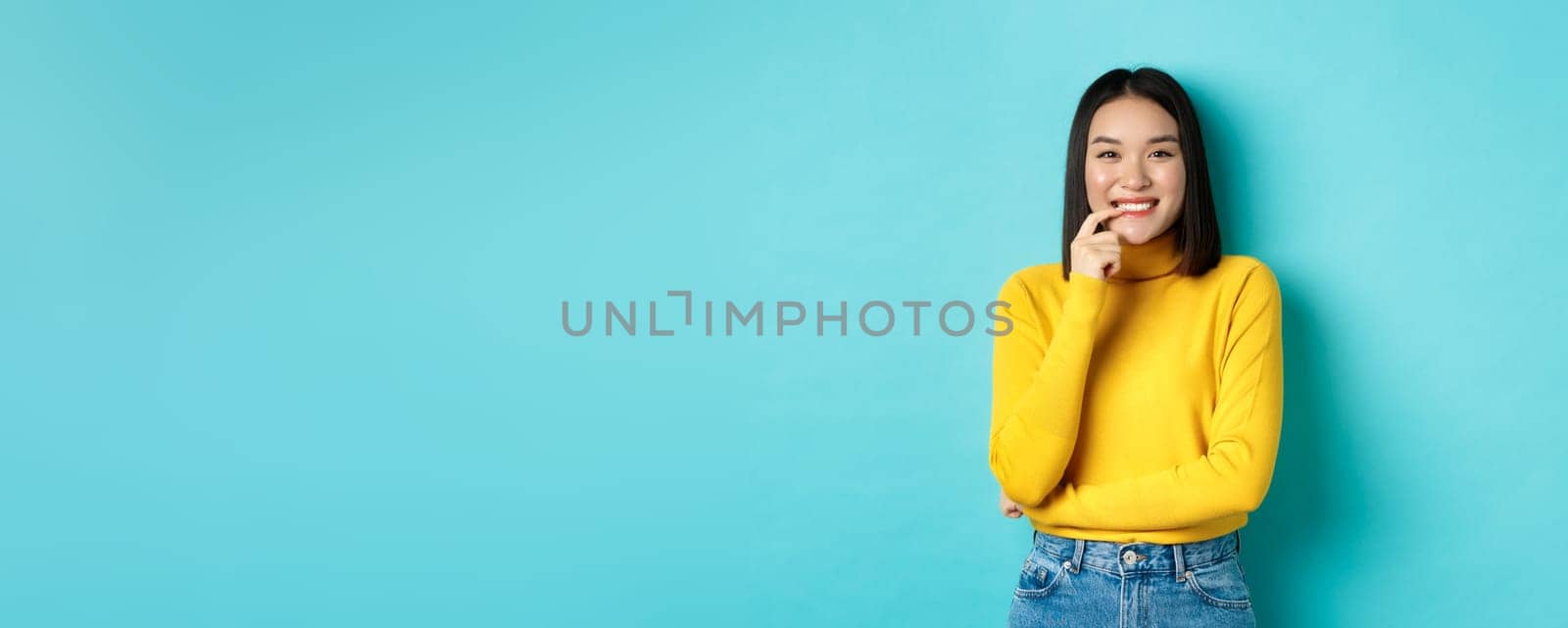 Image of good-looking asian woman in stylish outfit, touching lip and smiling at camera with happy face, standing over blue background by Benzoix