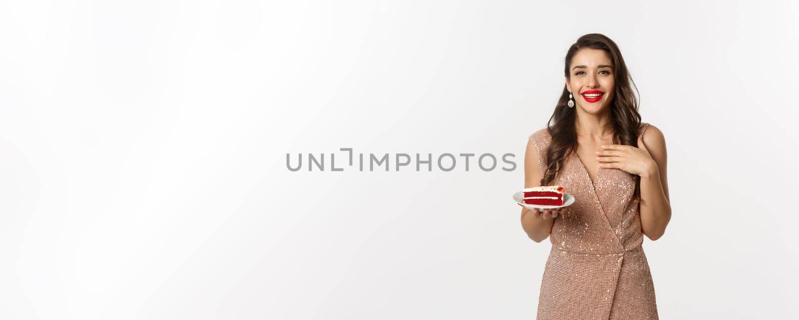 Party and celebration concept. Attractive slim woman in elegant dress holding piece of cake and smiling, standing over white background.