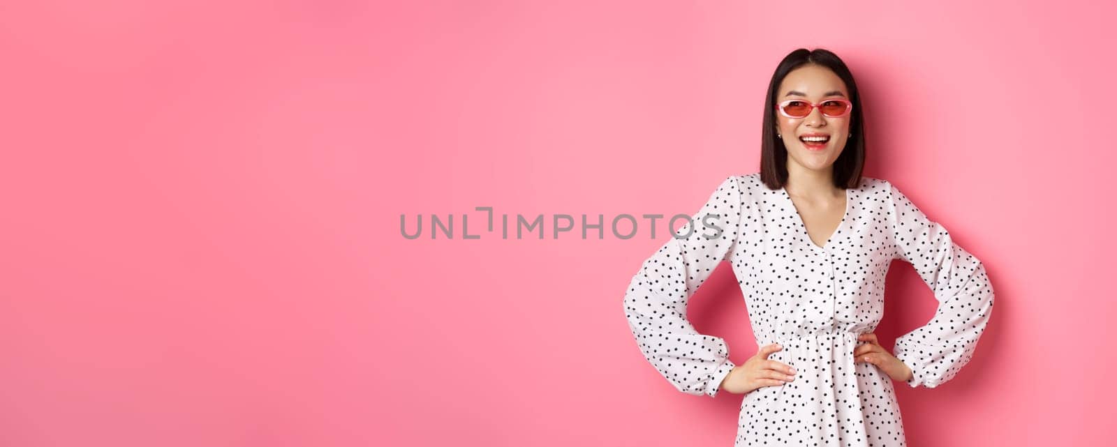 Tourism and lifestyle concept. Beautiful korean woman on vacation, wearing sunglasses and dress, looking left with happy smile, standing over pink background by Benzoix