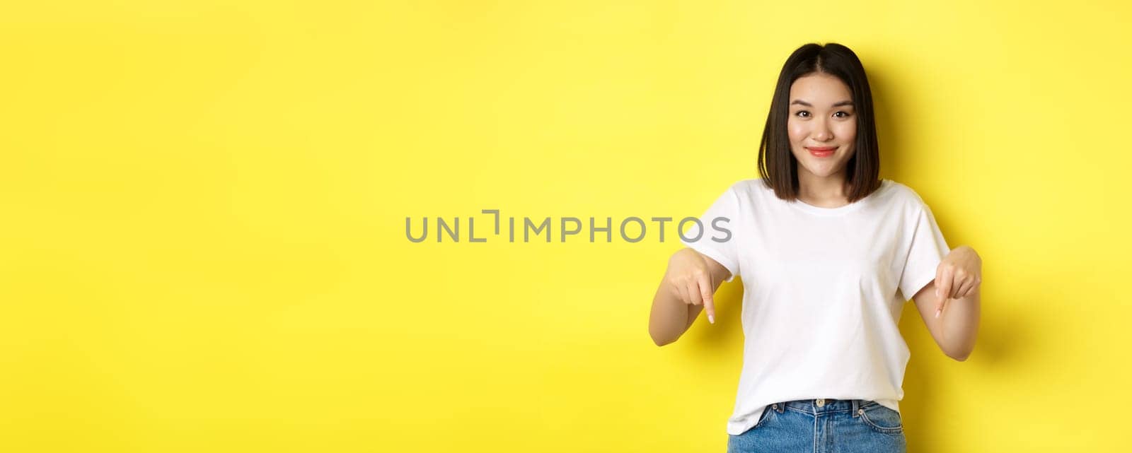 Beauty and fashion concept. Beautiful asian woman in white t-shirt pointing fingers down, demonstrate logo standing over yellow background by Benzoix