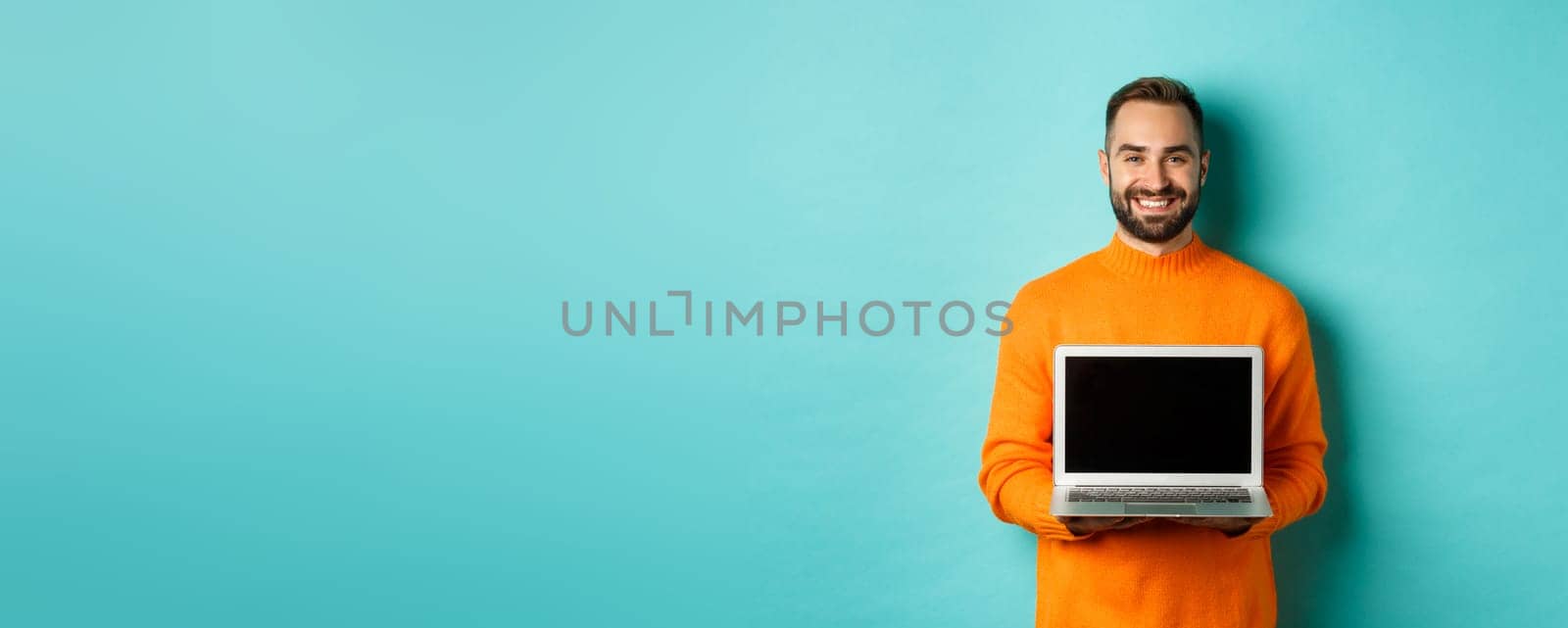 Handsome bearded man in orange sweater showing laptop screen, demonstrating online store, standing over light blue background by Benzoix