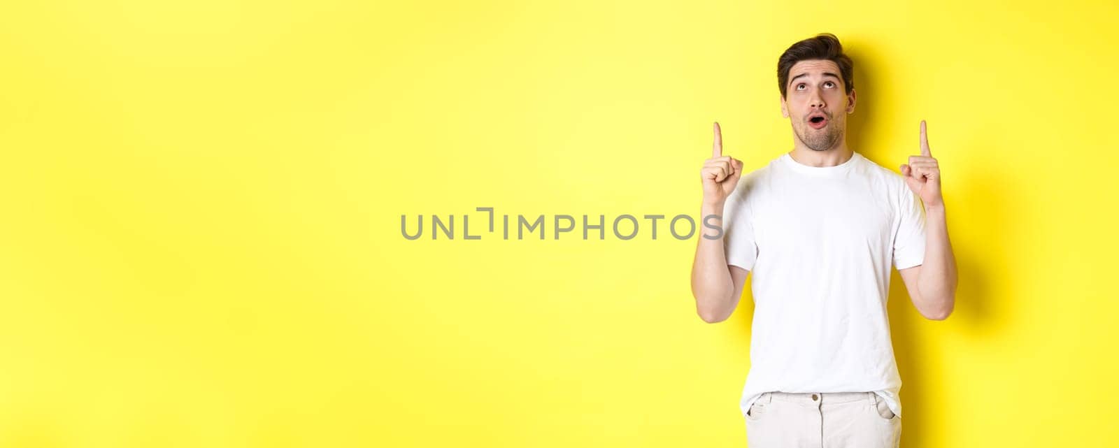 Impressed guy checking out promo offer, looking and pointing fingers up at advertisement, standing over yellow background by Benzoix