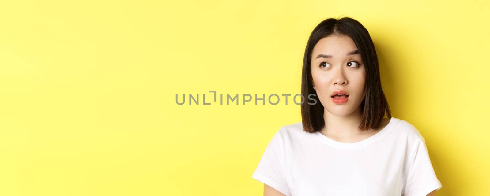 Close up of beautiful asian woman with clean perfect skin, raising eyebrow and looking left with intrigued face, standing over yellow background by Benzoix
