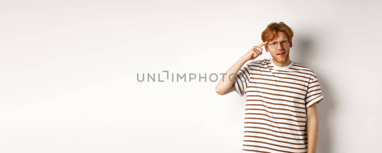 Annoyed young man with red hair scolding person for being stupid, pointing at head and staring at camera, white background.