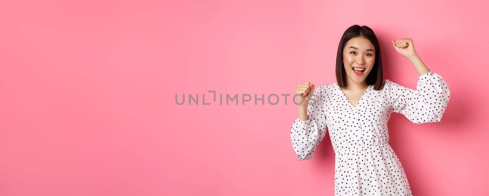 Beautiful korean woman dancing and having fun, smiling happy at camera, posing against pink background.