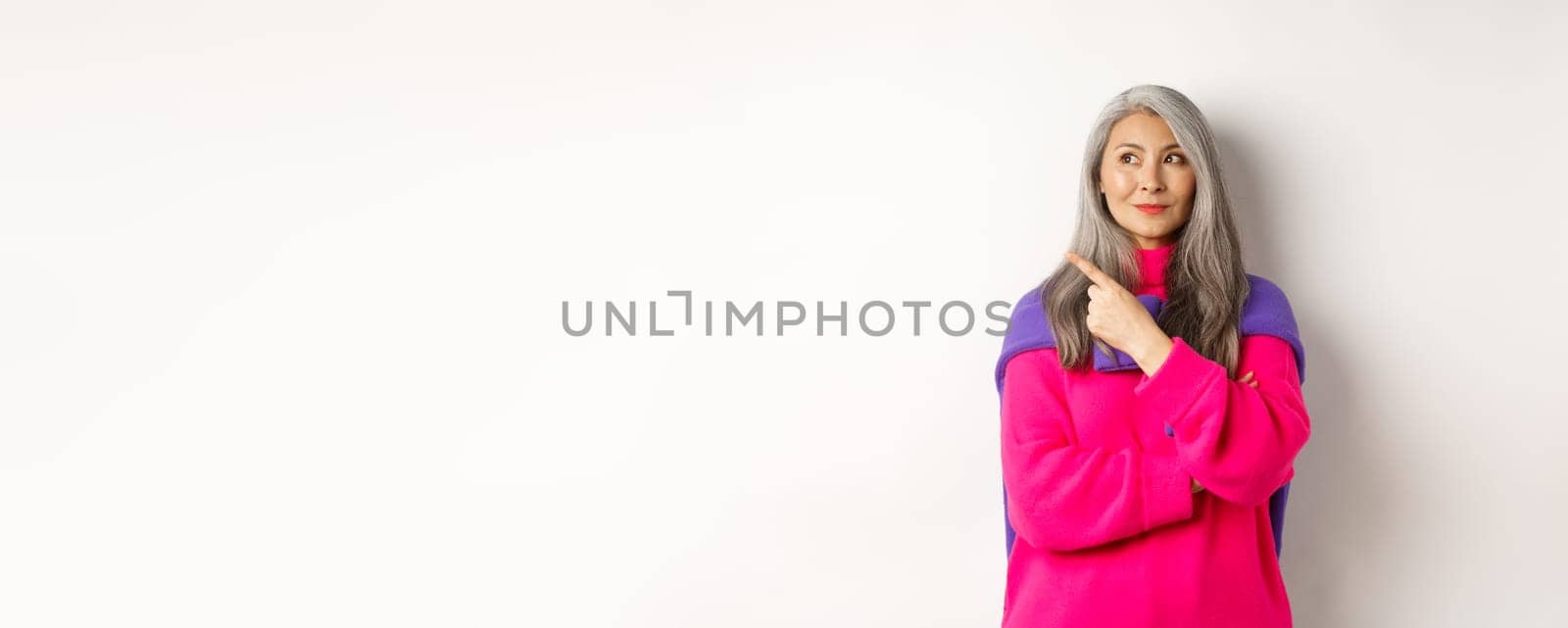 Stylish modern asian elderly woman pointing at upper left corner, looking at logo with pleased smile, standing in pink sweater over white background.