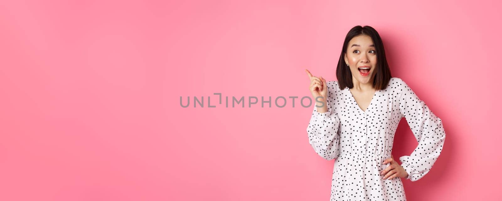 Excited asian woman in stylish dress, gasping amused, pointing finger upper left corner, showing interesting promo offer, standing over pink background by Benzoix