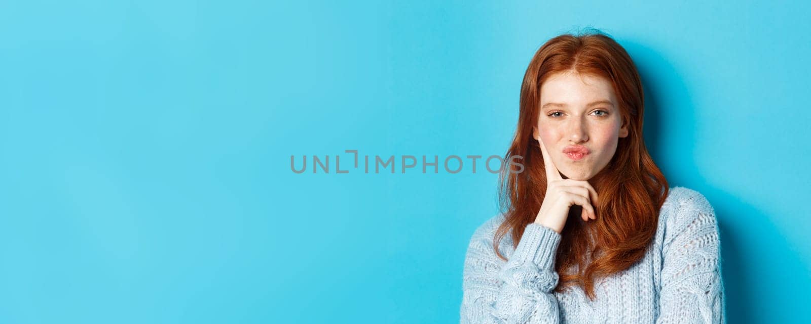 Close-up of beautiful redhead girl thinking, pucker lips and staring thoughtfula t camera, making choice, standing over blue background.