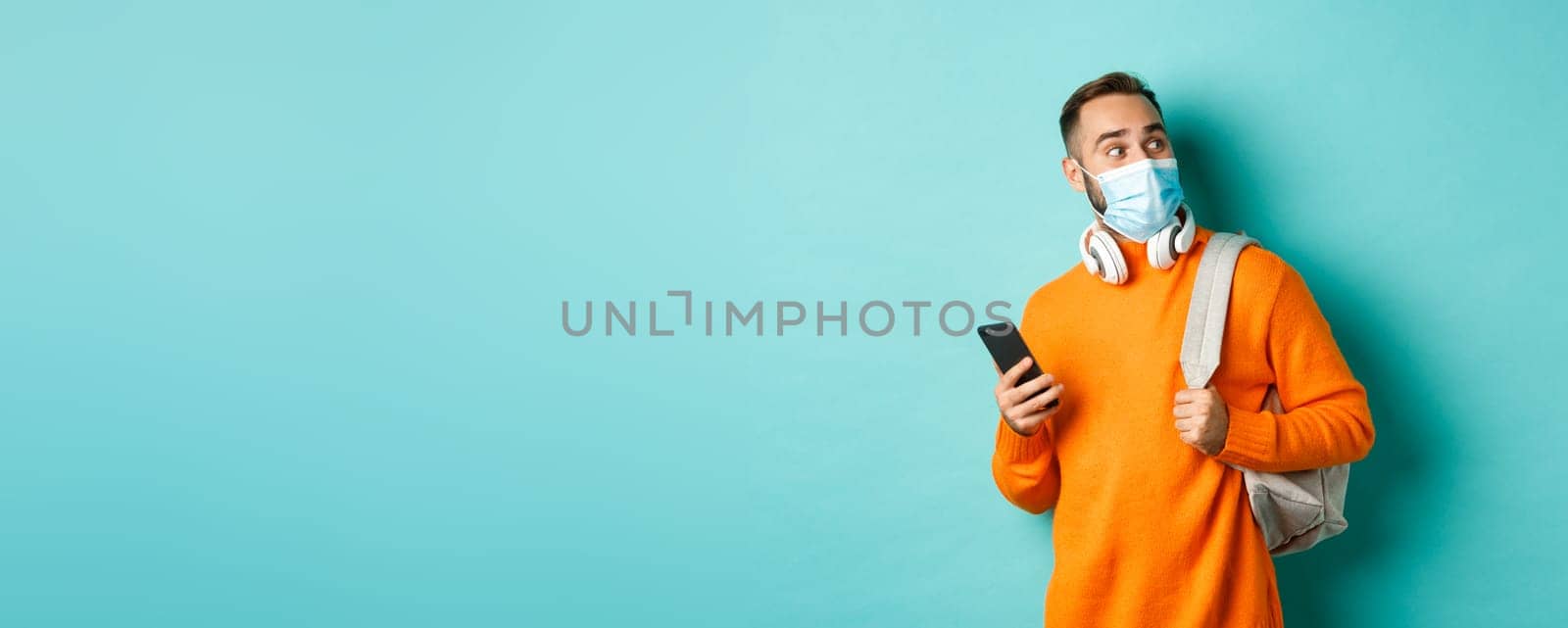 Young man in face mask using mobile phone, holding backpack, staring right amazed, standing against light blue background.