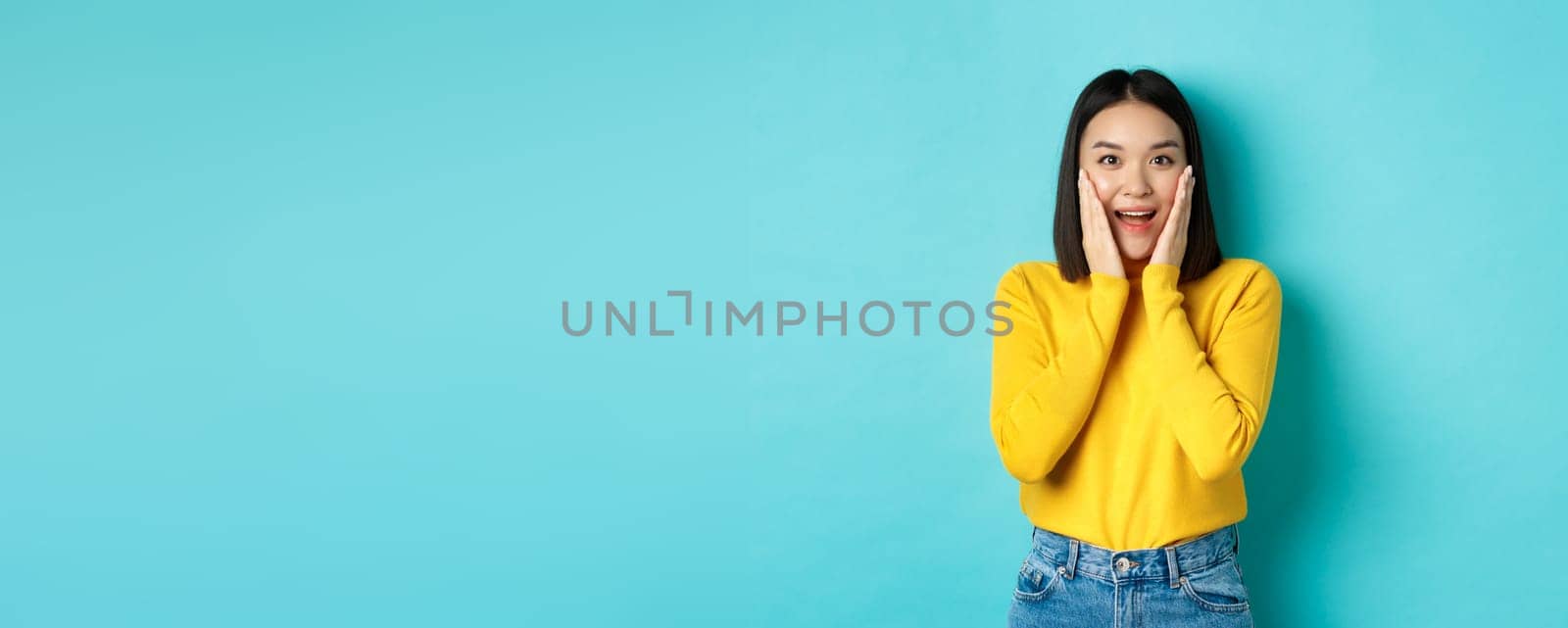 Image of cheerful and surprised asian woman checking out promotion, gasping amazed, standing over blue background by Benzoix