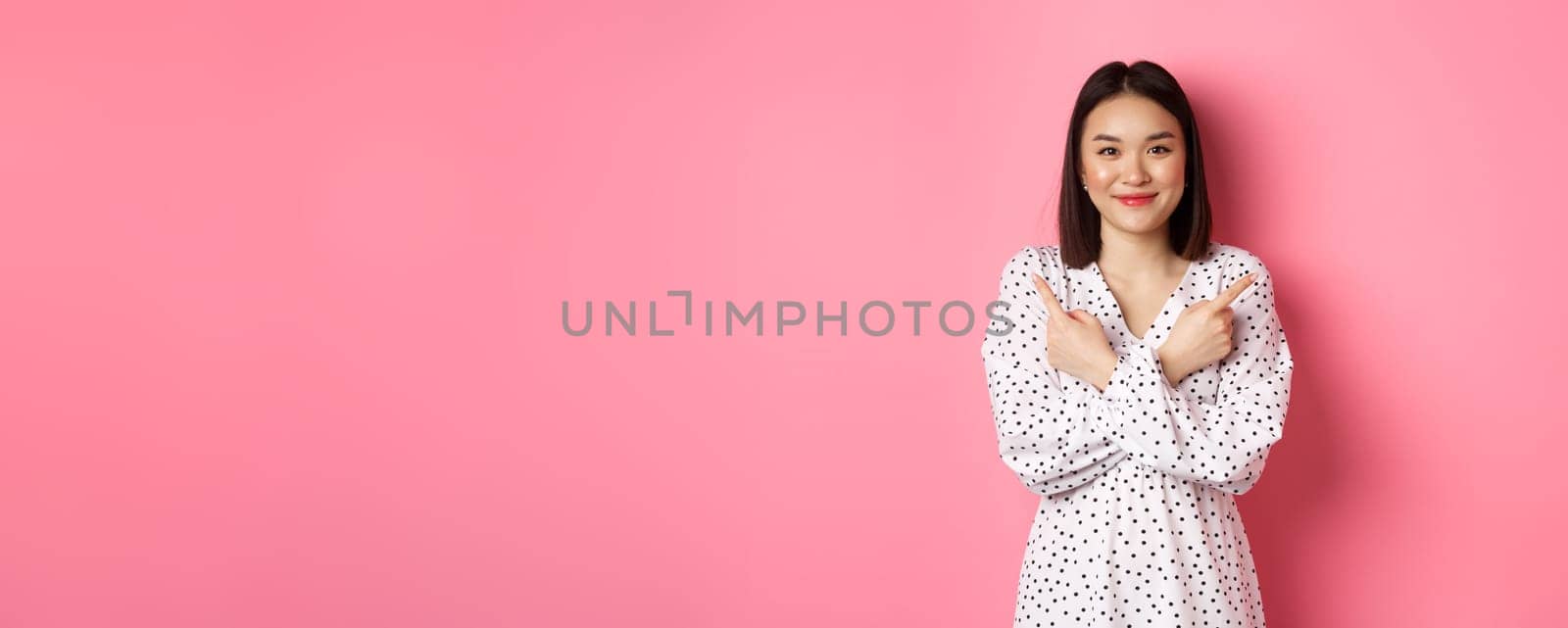 Beautiful asian girl in romantic dress pointing fingers sideways, showing two variants on shopping, smiling at camera, standing against pink background by Benzoix