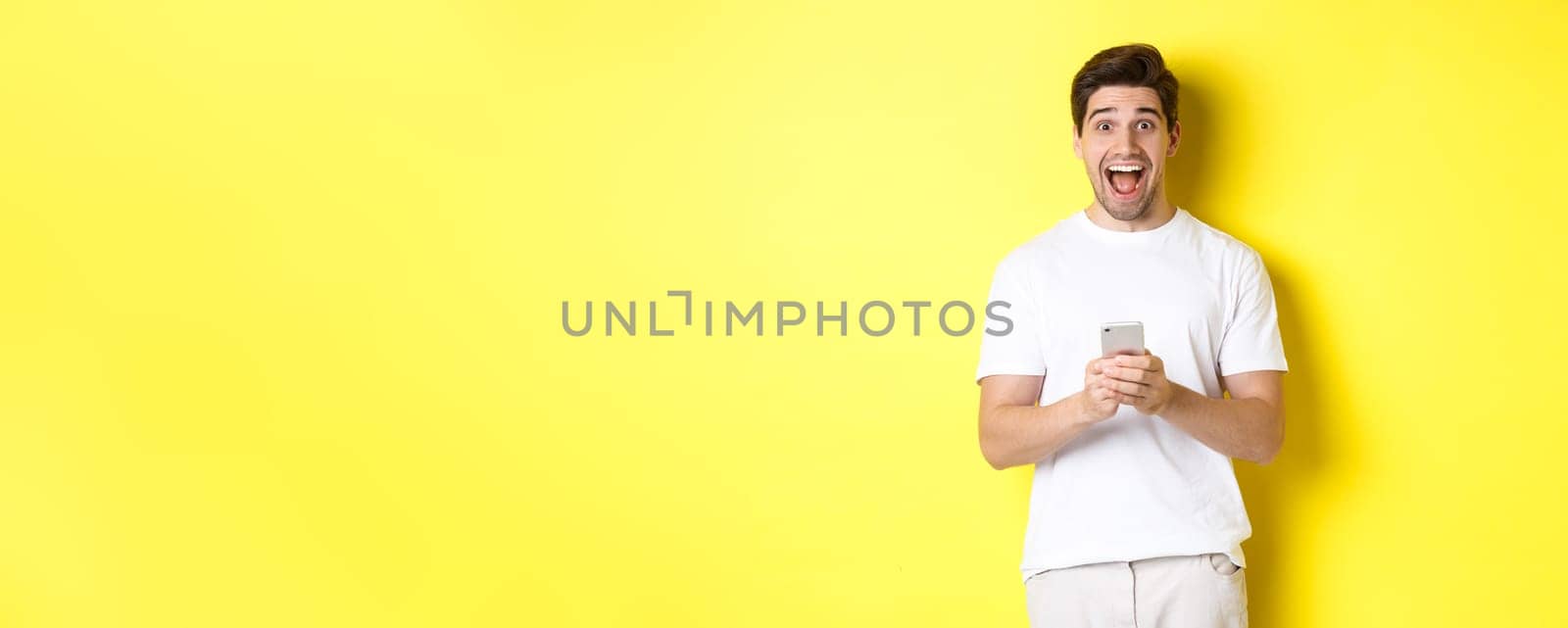 Man looking amazed and happy after reading something on mobile phone, standing over yellow background. Copy space