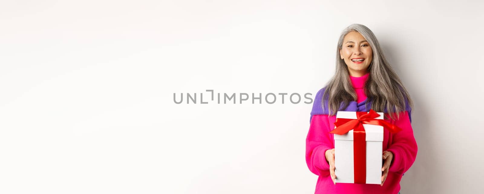 Beautiful asian senior woman smiling, congratulating with valentines day, holding gift in box, standing over white background by Benzoix