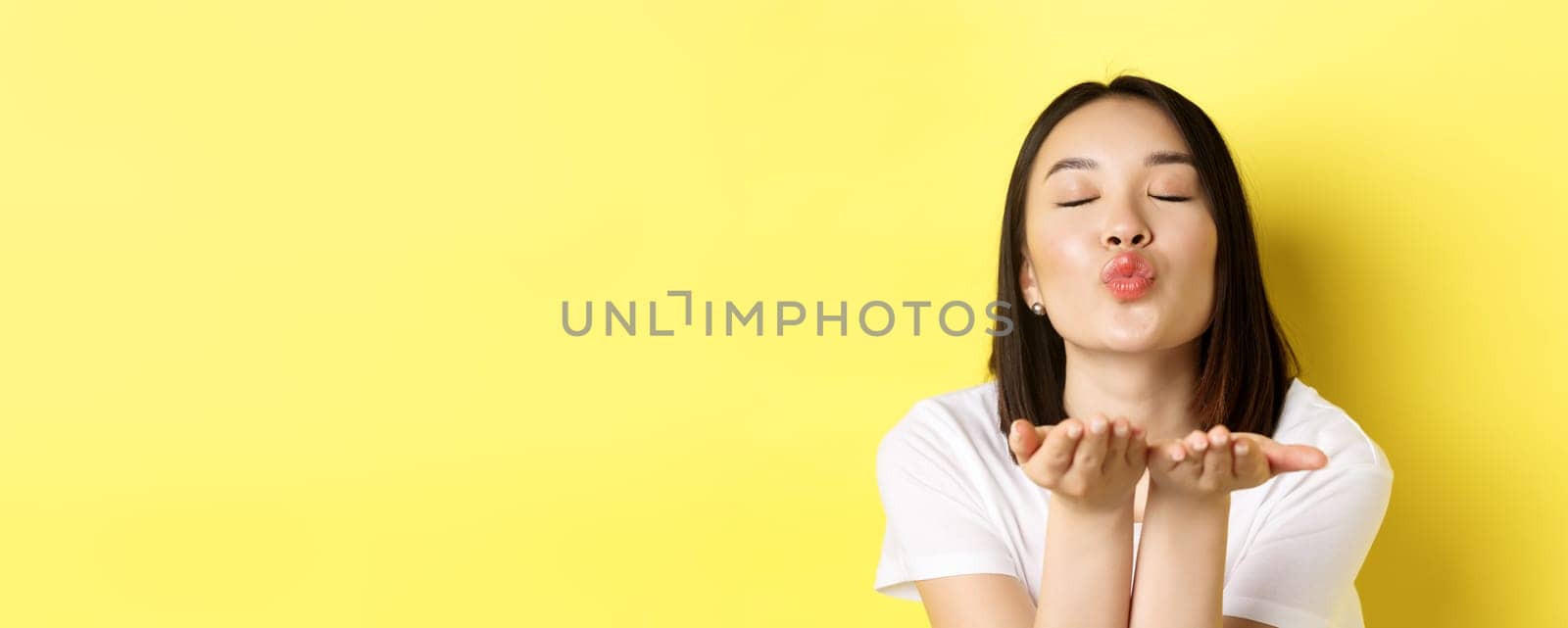 Valentines day concept. Close up of beautiful asian woman pucker lips and holding hands near mouth, blow air kiss at camera with closed eyes, standing over yellow background.