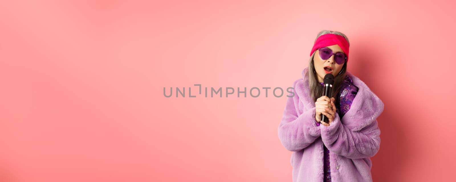 Fashion concept. Stylish senior asian woman singing karaoke, perform on stage with microphone, wearing trendy sunglasses and purple faux fur coat, pink background by Benzoix