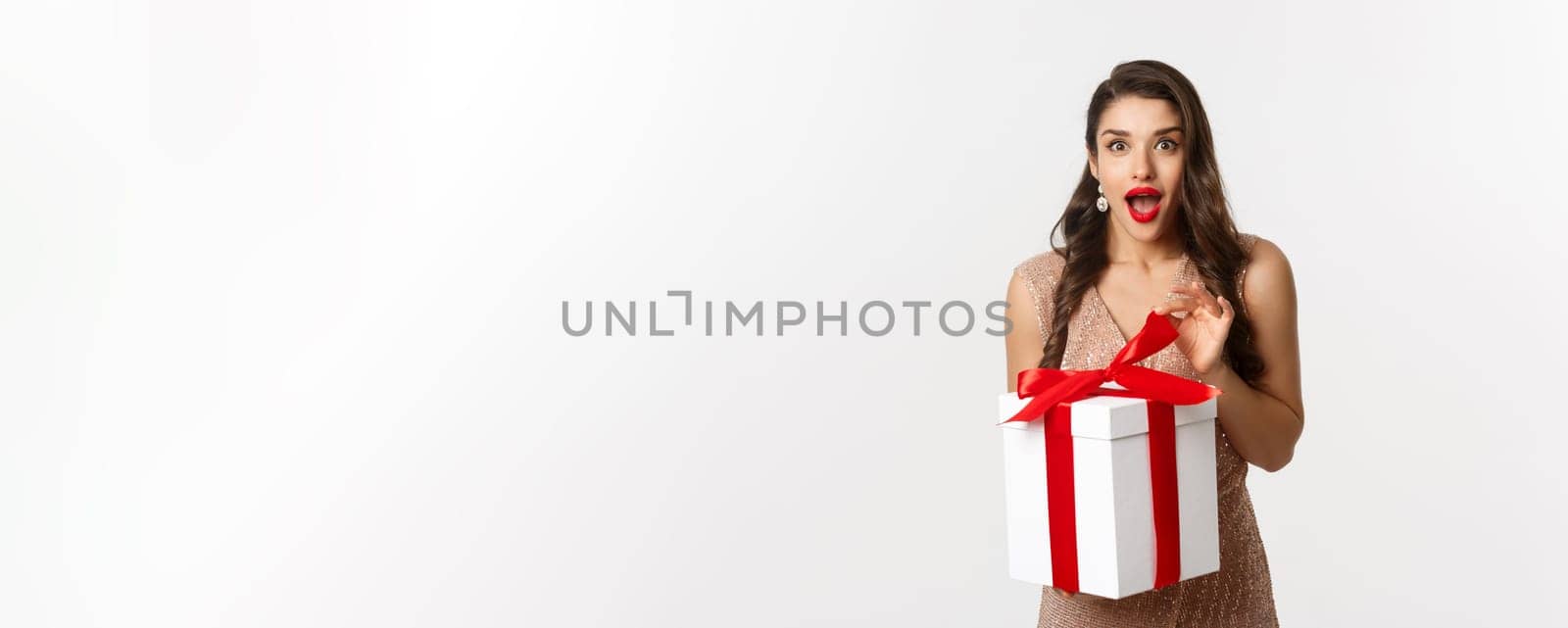 Merry Christmas. Image of attractive woman in luxury dress, open gift and looking surprised, receive new year present, standing over white background by Benzoix