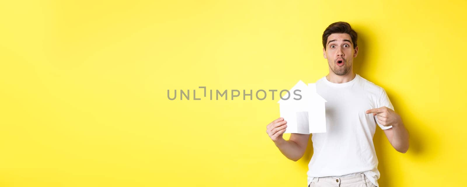 Real estate concept. Excited man pointing finger at paper house model, searching for flat, standing over yellow background by Benzoix