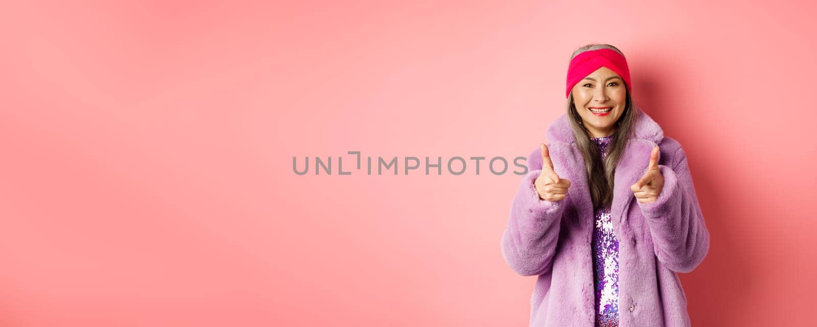 Fashion and shopping concept. Cool asian senior woman in stylish fake fur coat pointing fingers at camera, asking you to check out promo offer, standing over pink background.