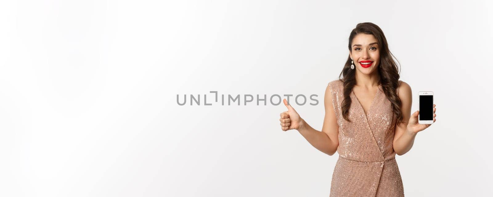 Online shopping. Beautiful young woman in party dress and makeup, showing thumbs up and mobile screen, praising good app, white background.