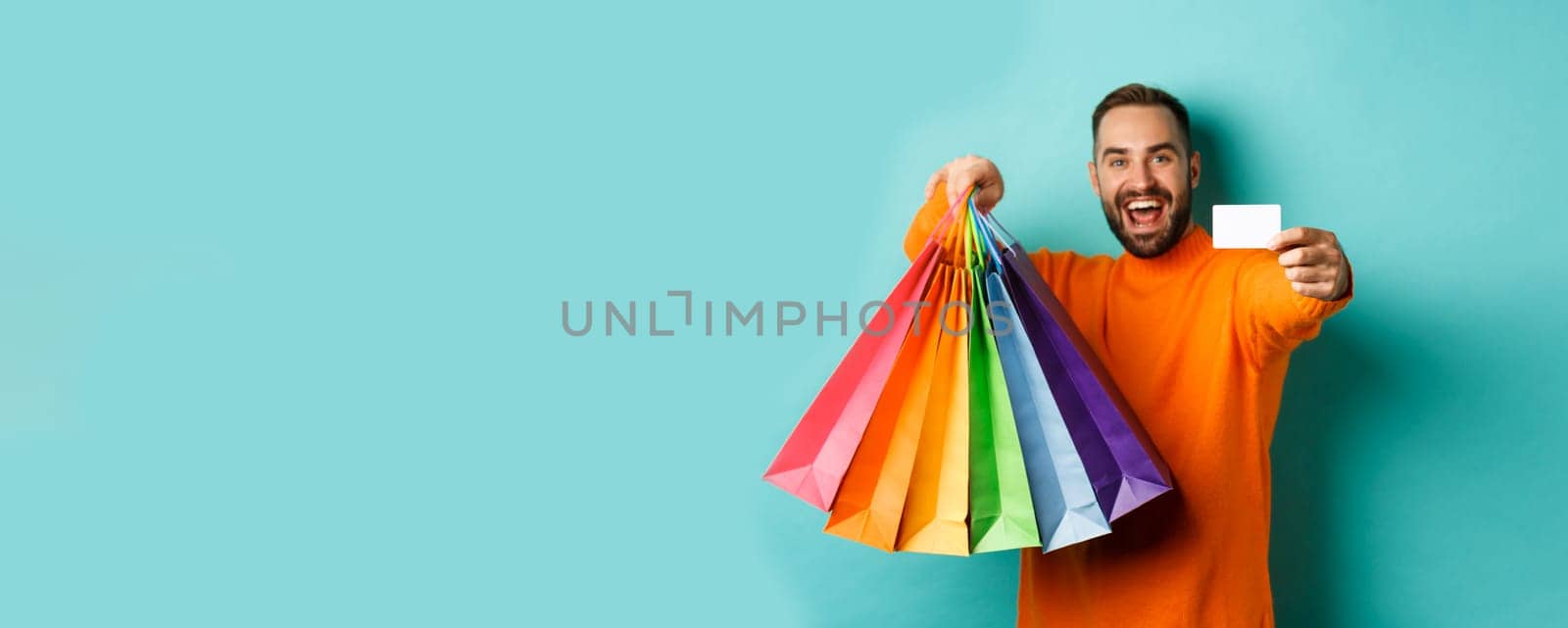 Happy aduly man showing credit card and shopping bags, standing against turquoise background by Benzoix