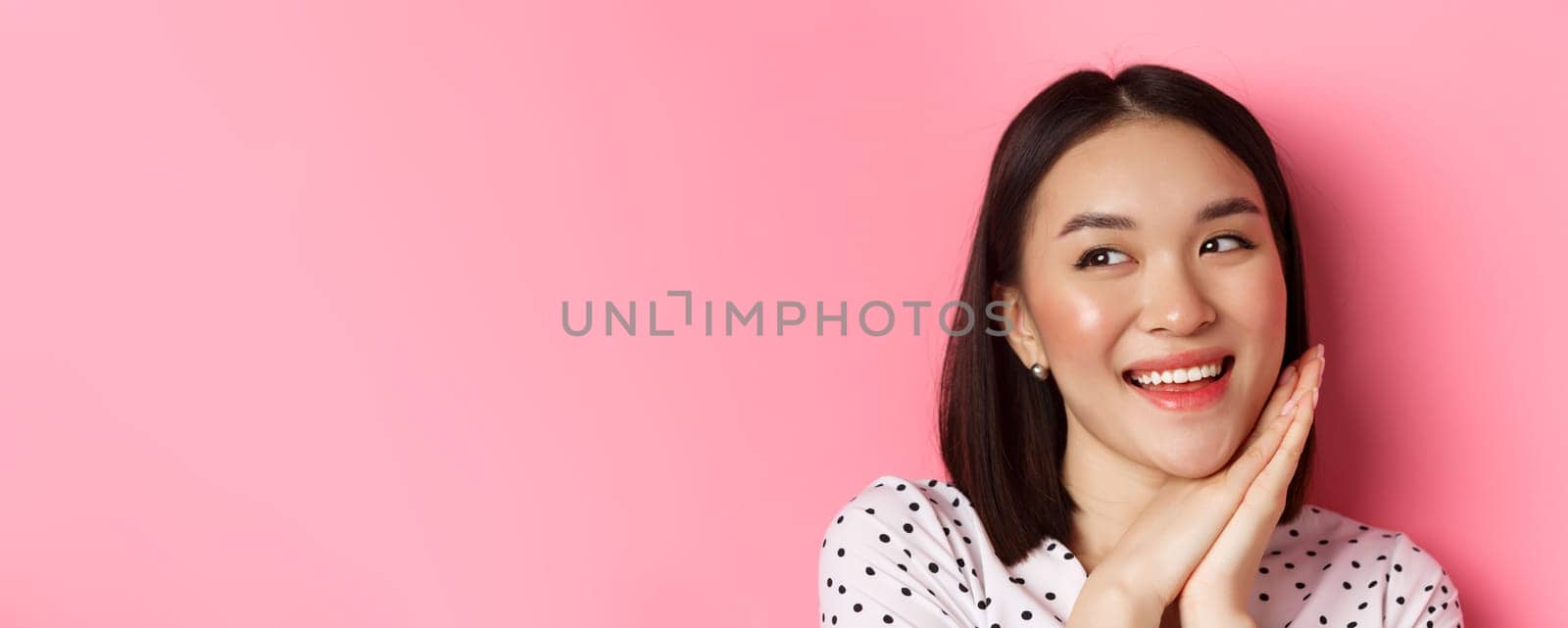 Beauty and skin care concept. Headshot of adorable and dreamy asian woman looking left, smiling and imaging, standing against pink background.