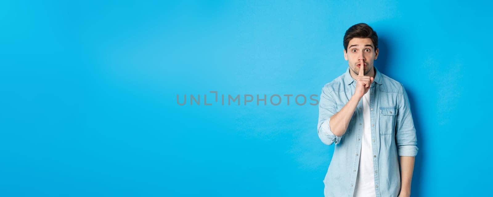 Portrait of excited man asking to keep quiet, showing hush taboo sign and looking nervously at camera, standing against blue background.