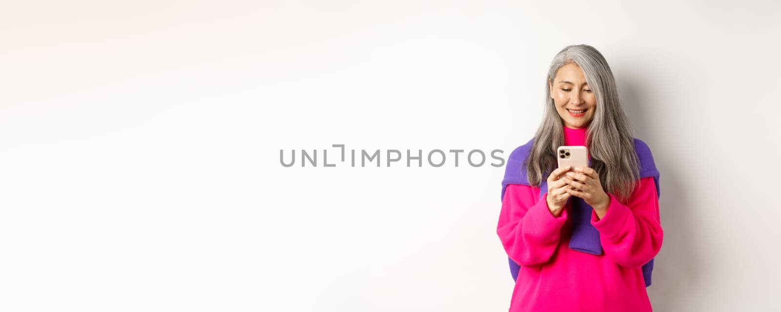 Beautiful asian senior woman in pink stylish sweater using smartphone. Old lady reading on mobile phone and smiling, standing over white background.