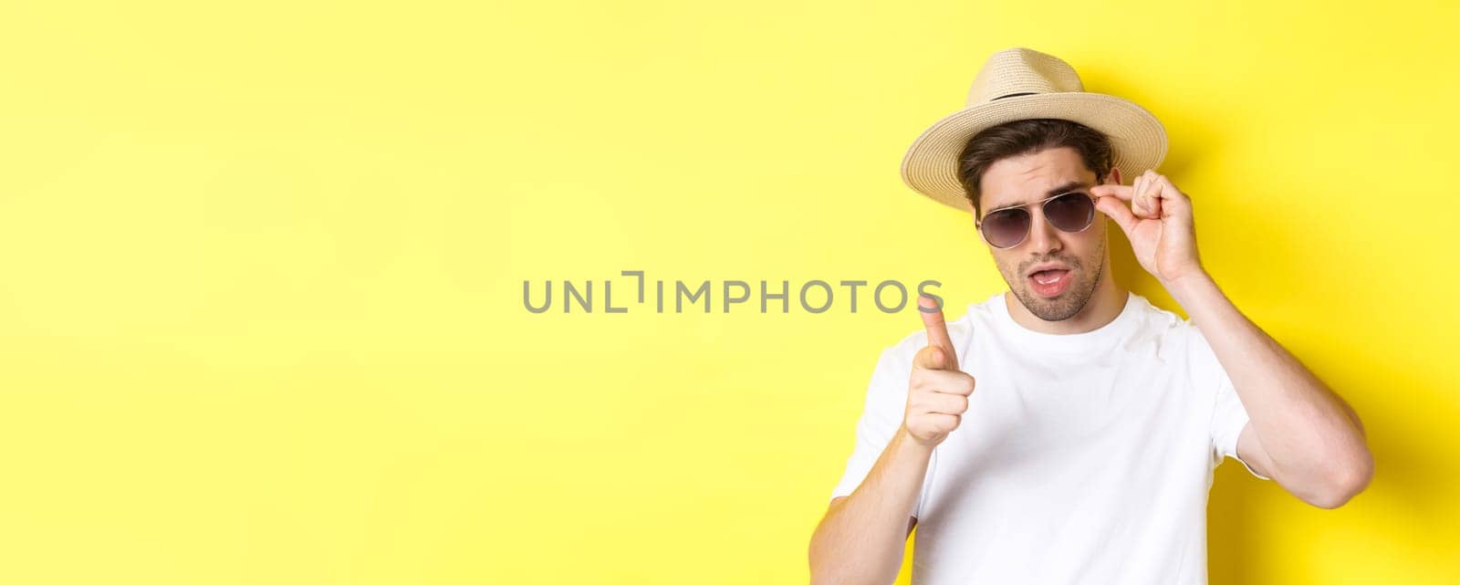 Concept of tourism and vacation. Cool and sassy man flirting with you, wearing sunglasses and pointing finger at camera, standing over yellow background.
