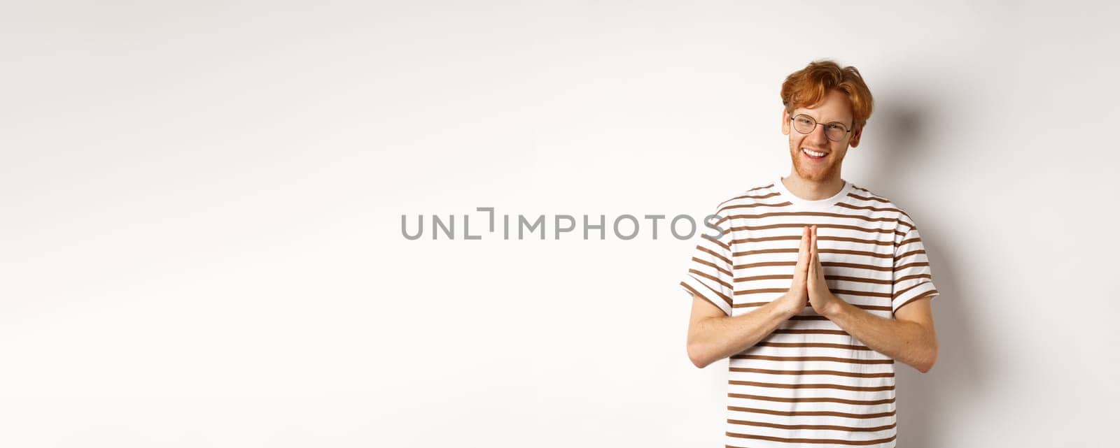 Handsome young man in glasses, red hair, showing namaste gesture and smiling, thanking you, standing grateful over white background.