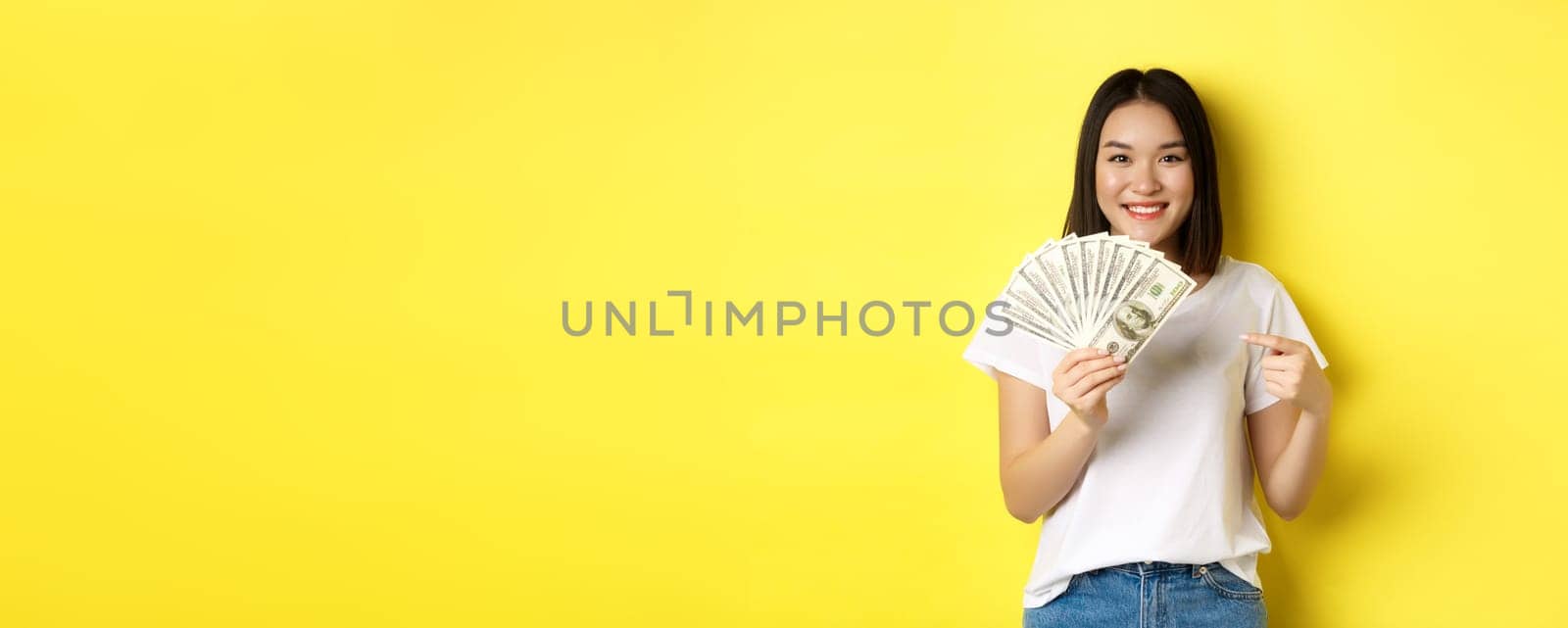 Young asian woman smiling, showing prize money, pointing finger at dollars, standing over yellow background by Benzoix