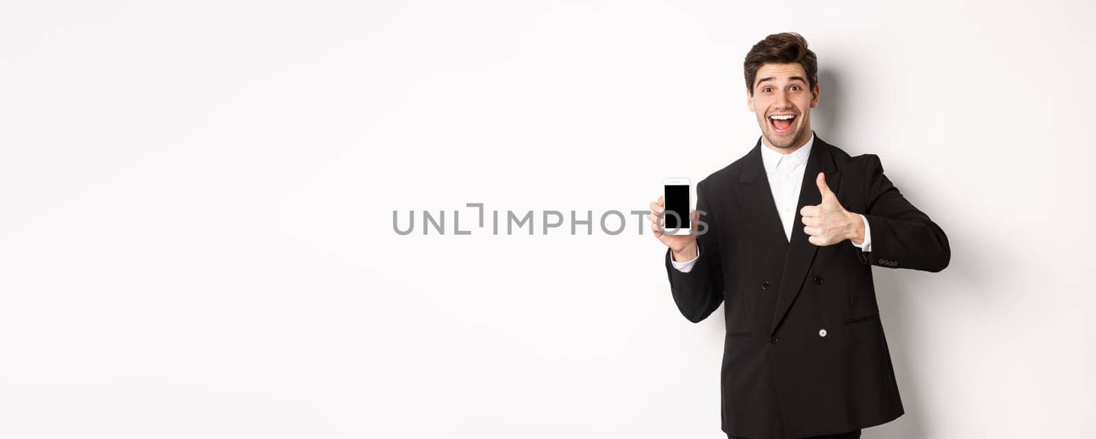 Image of handsome male entrepreneur in black suit, recommending app or online shop, showing thumbs-up and smartphone screen, standing over white background.