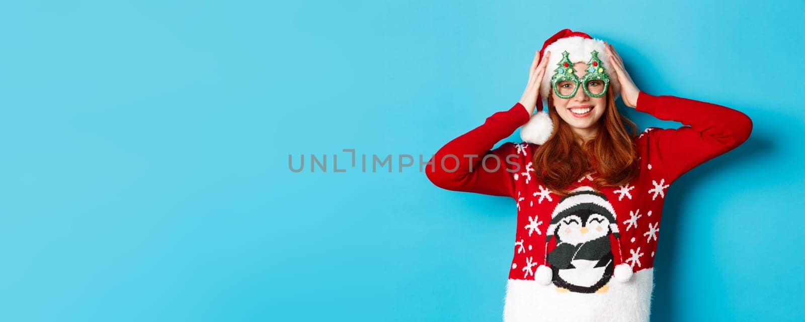 Happy holidays and Christmas concept. Funny redhead teen girl celebrating New Year, wearing santa hat and party glasses, standing against blue background by Benzoix