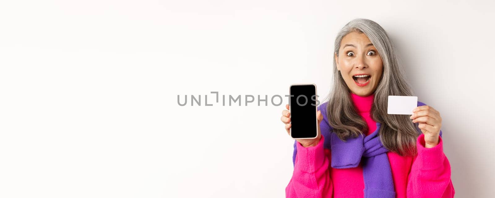 Online shopping. Closeup of amazed asian senior woman showing blank mobile screen and plastic credit card, looking happy, standing over white background.