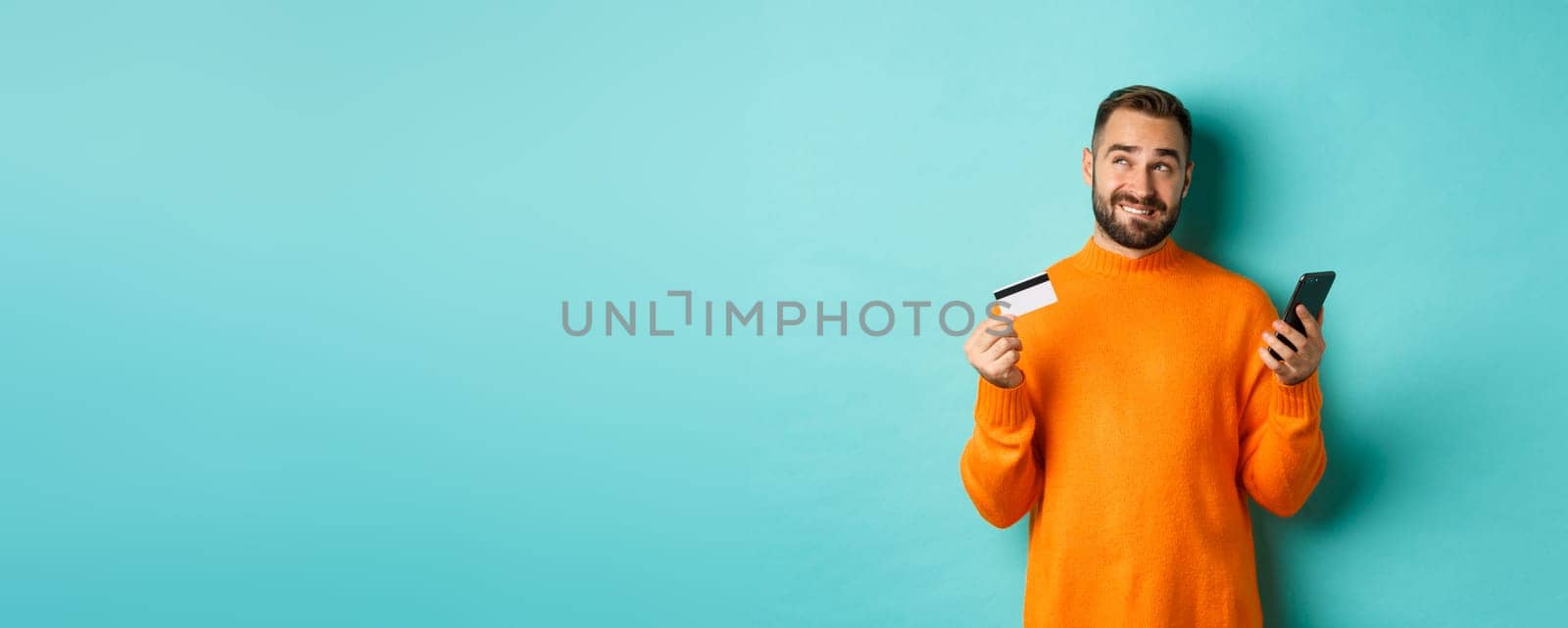 Online shopping. Thoughtful man holding credit card and mobile phone, thinking of purchase, standing over light blue background.