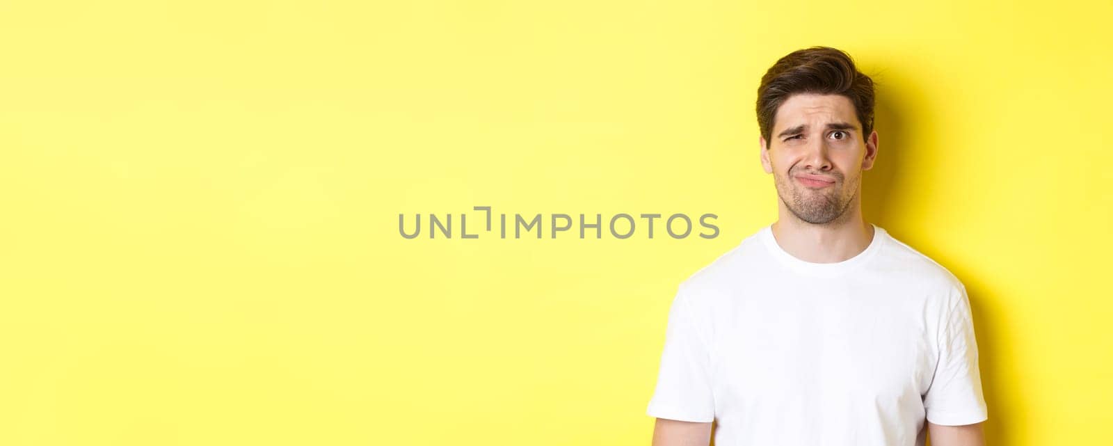 Close-up of dispelased young man in white t-shirt looking doubtful, grimacing unsatisfied, standing over yellow background.