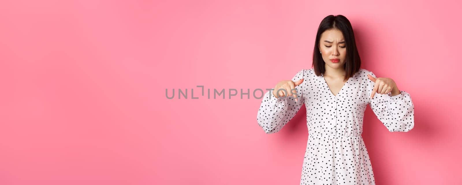 Concerned asian woman peeking down and pointing hand at disturbing banner, frowning upset, standing over pink background.