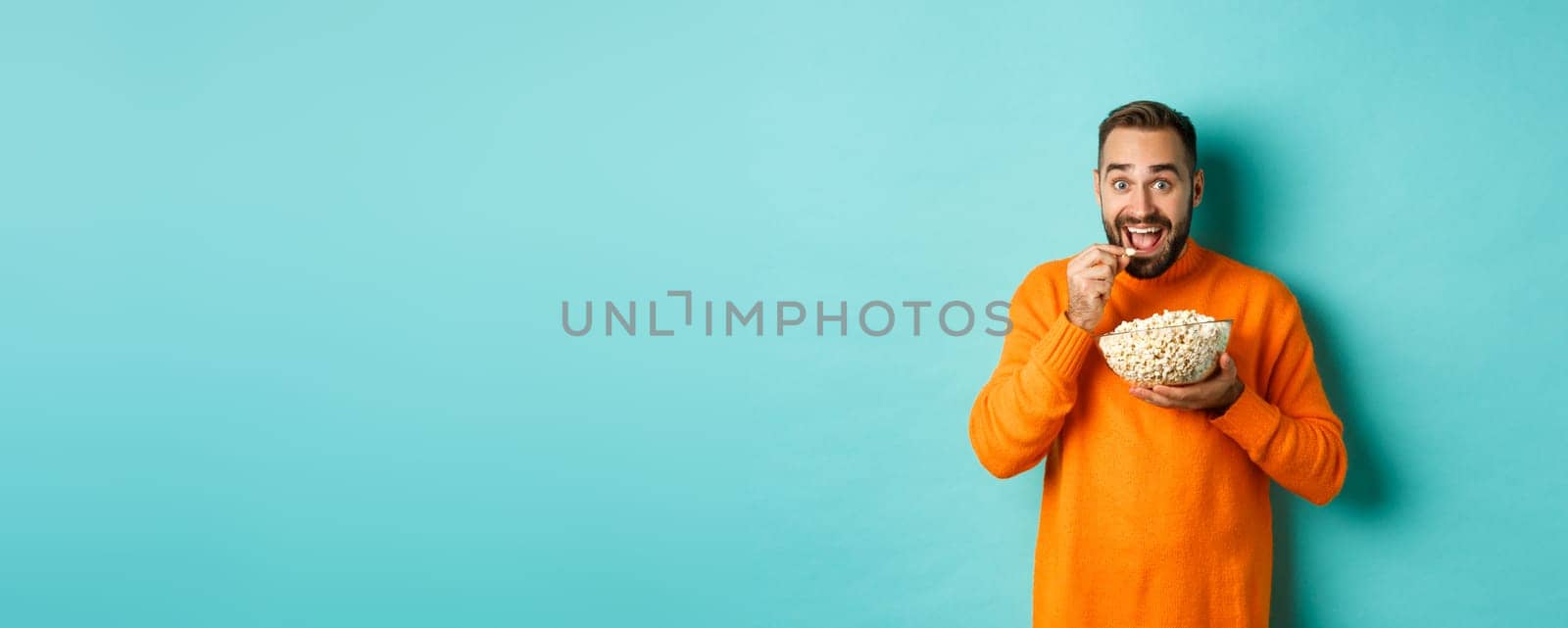 Excited young man watching interesting movie on tv screen, eating popcorn and looking amazed, blue background.