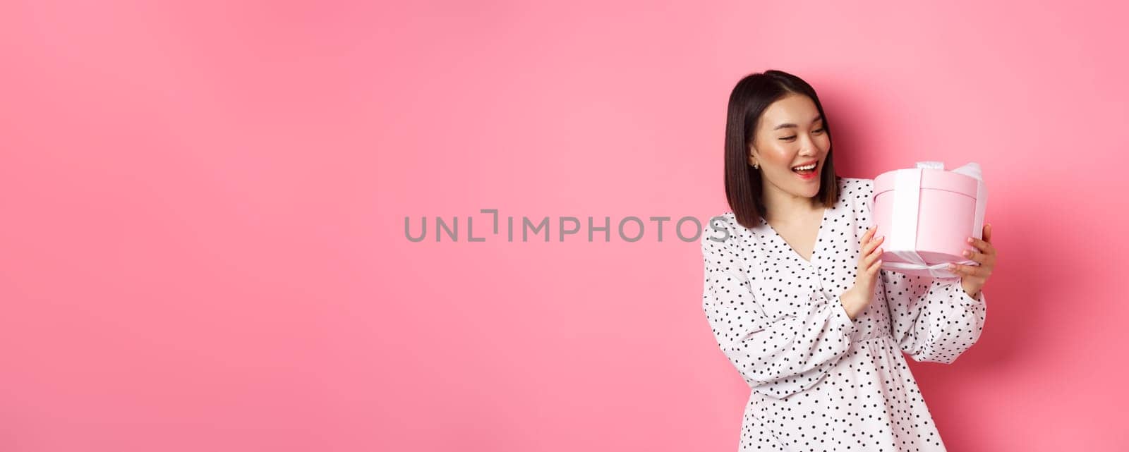 Valentines day, celebration concept. Beautiful asian woman holding romantic gift box, smiling happy, standing over pink background by Benzoix