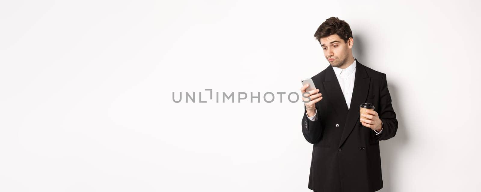 Portrait of thoughtful handsome businessman, drinking coffee and browsing in internet, looking at smartphone screen, standing against white background.